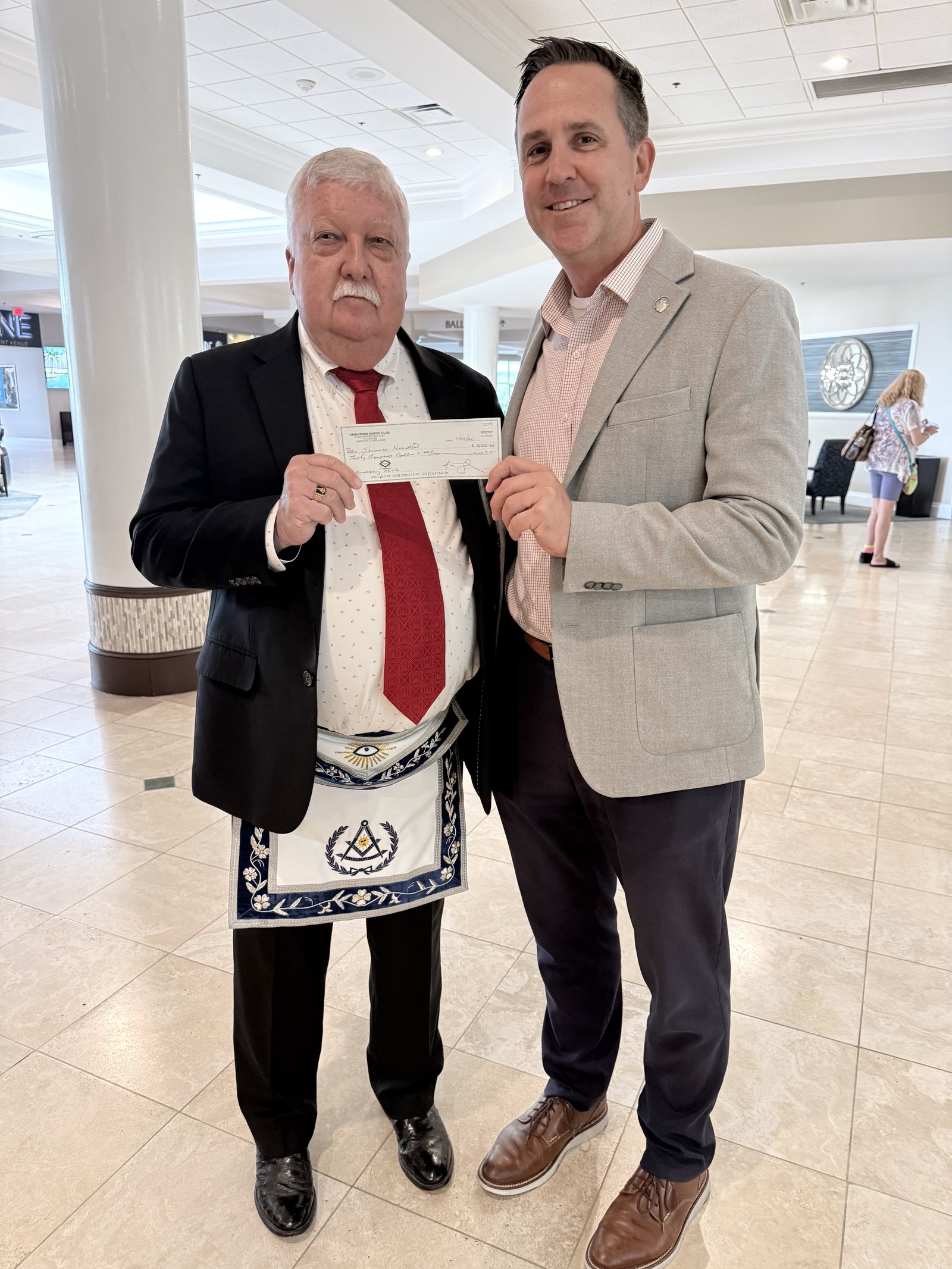 Two Marathon Shrine Club members holding a presentation check inside the lobby of Shriners Children's, Tampa.