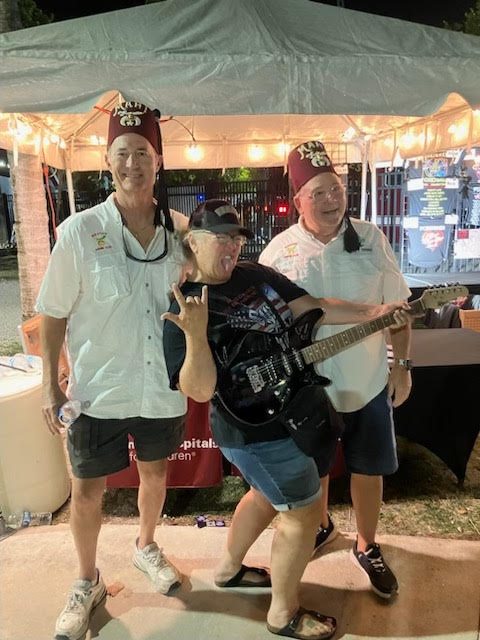 The winner of the signed Foreigner guitar holding it under the raffle tent, flanked by two Shriners in fezzes.